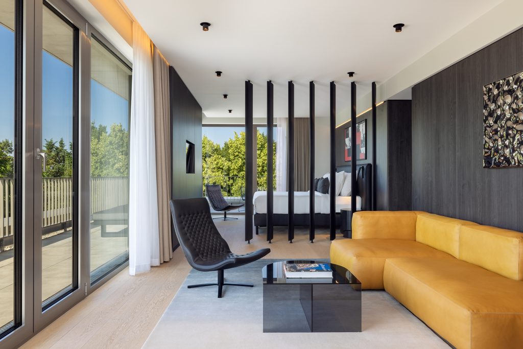 Moderne Hotel-Suite mit offener Raumaufteilung, bodentiefen Fenstern, Designermöbeln und gelbem Ledersofa.