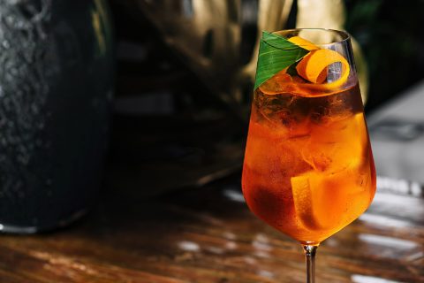 Cocktailglas steht mit Eis und Orangenscheibe auf rustikalem Holztisch