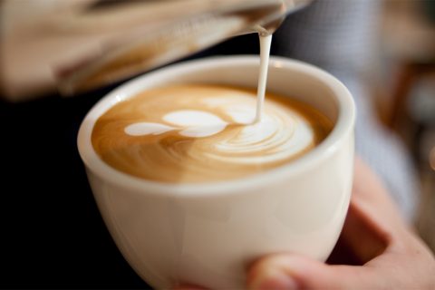 Tasse Cappuccino: Milch wird zu Herz-Latteart gegossen, Hand hält Tasse