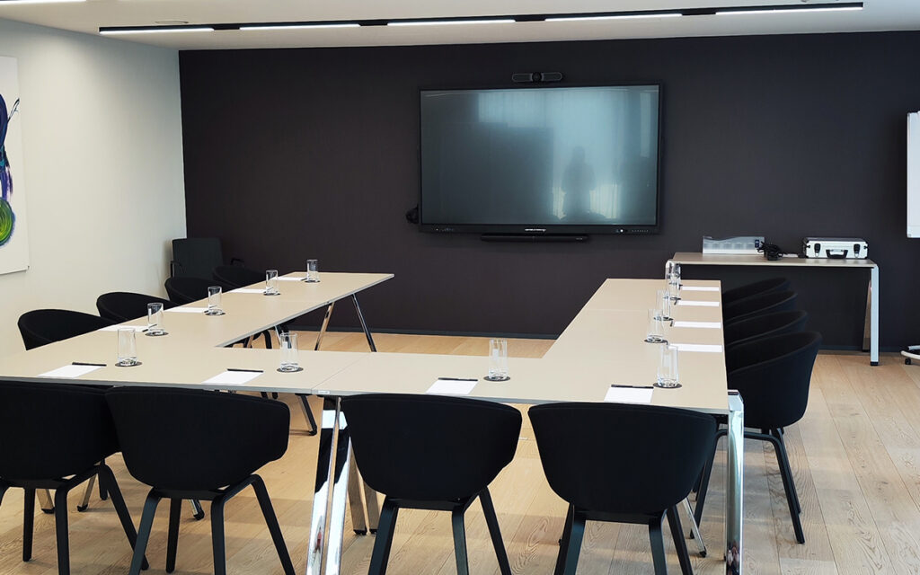 U-förmiger Konferenztisch, bestückt mit Wassergläsern und Notizblättern, bereit für eine Besprechung, in modernem Besprechungsraum in Stuttgart mit großem Wandmonitor, schwarzer Rückwand, Holzfußboden und Stühlen.