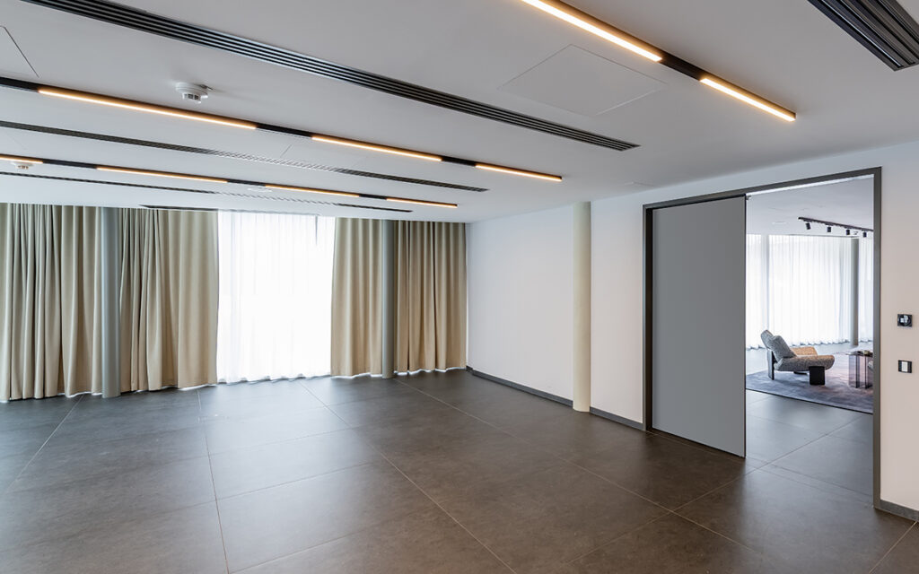 Großer, leerer Veranstaltungsraum im Tagungsotel nahe Stuttgart— moderner Innenraum mit dunklem Fliesenboden, linearen Deckenleuchten und beigen Vorhängen; geöffnete Schiebetür führt zu möbliertem Lounge-Bereich.
