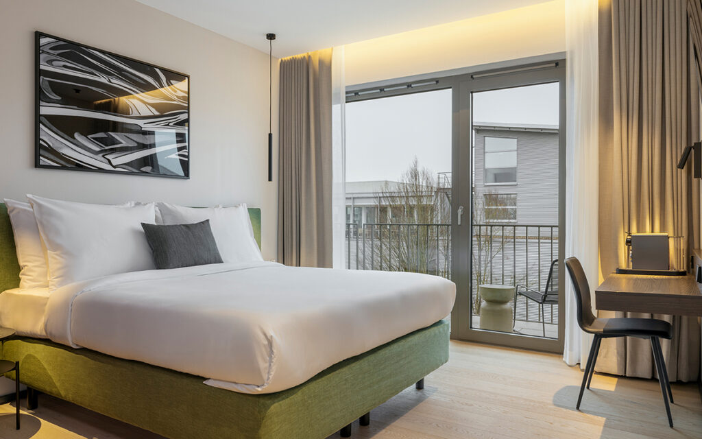 Weißes Bett, ordentlich bezogen, in einem modernen Hotelzimmer nahe Stuttgart mit bodentiefen Fenstern und kleinem Balkon, Schreibtisch, Stuhl und abstraktem Wandbild.
