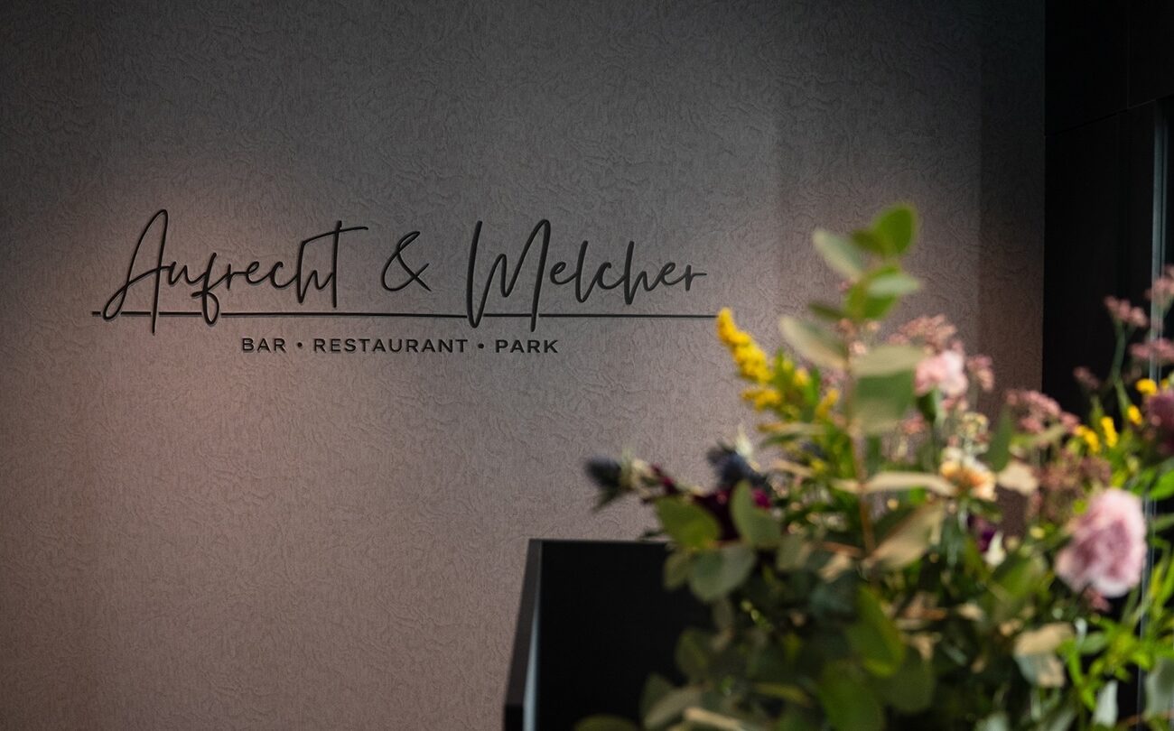 Wandlogo vor Blumen zeigt 'Aufrecht & Melcher' — BAR • RESTAURANT • PARK