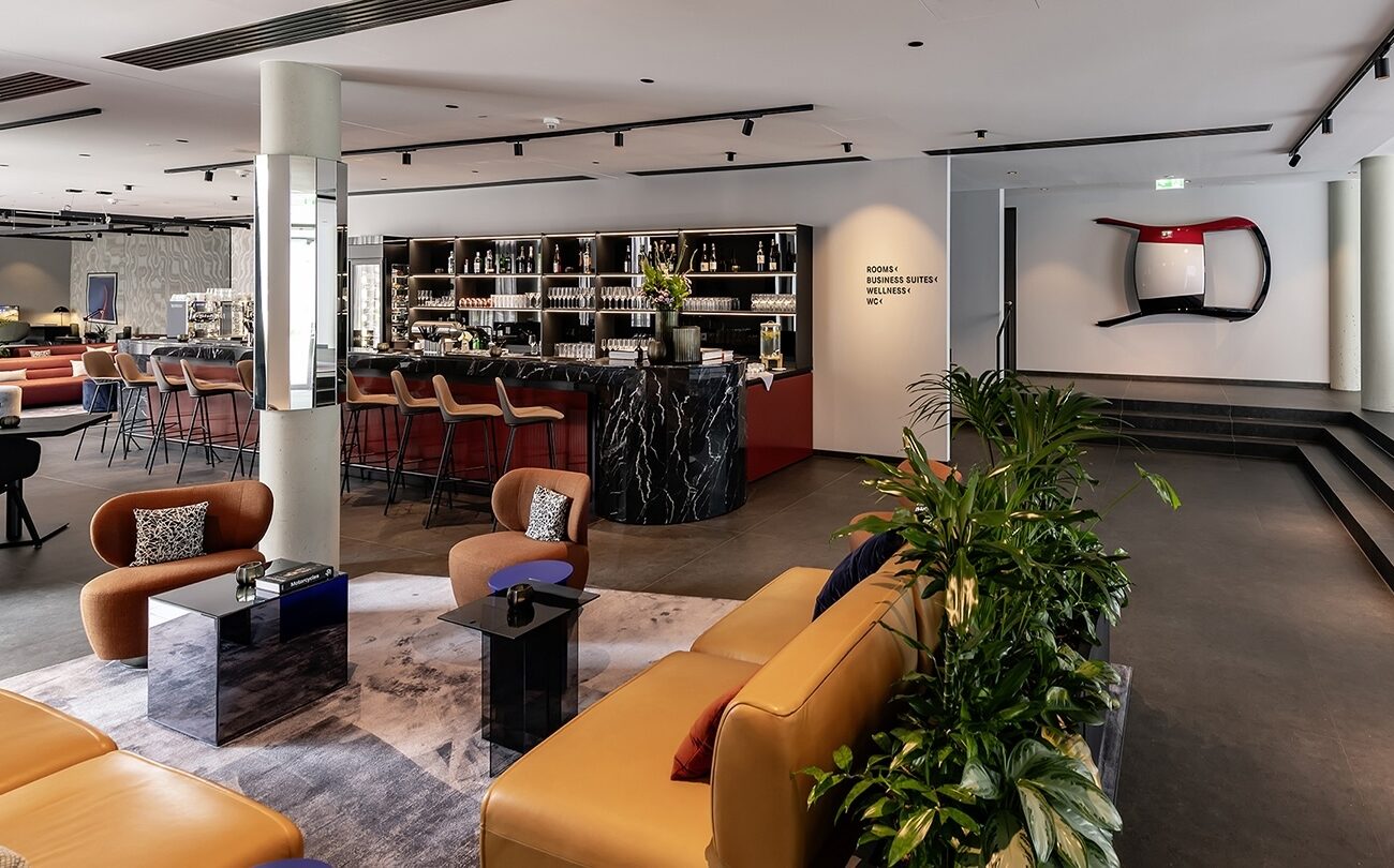 Hotelbar mit Sofas und Pflanzen; Text: ROOMS< BUSINESS SUITES< WELLNESS< WC<