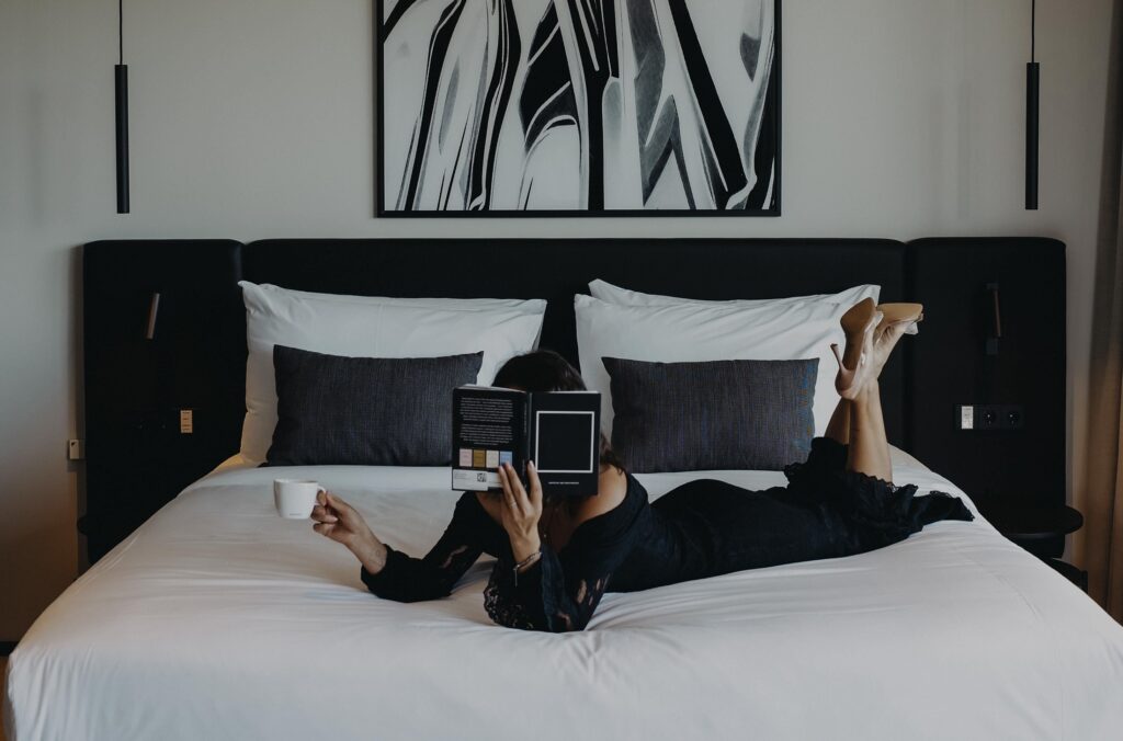 Eine Frau liegt auf dem Bauch auf einem großen weißen Bett, liest ein Buch, hält eine Kaffeetasse, Beine hoch mit Absatzschuhen